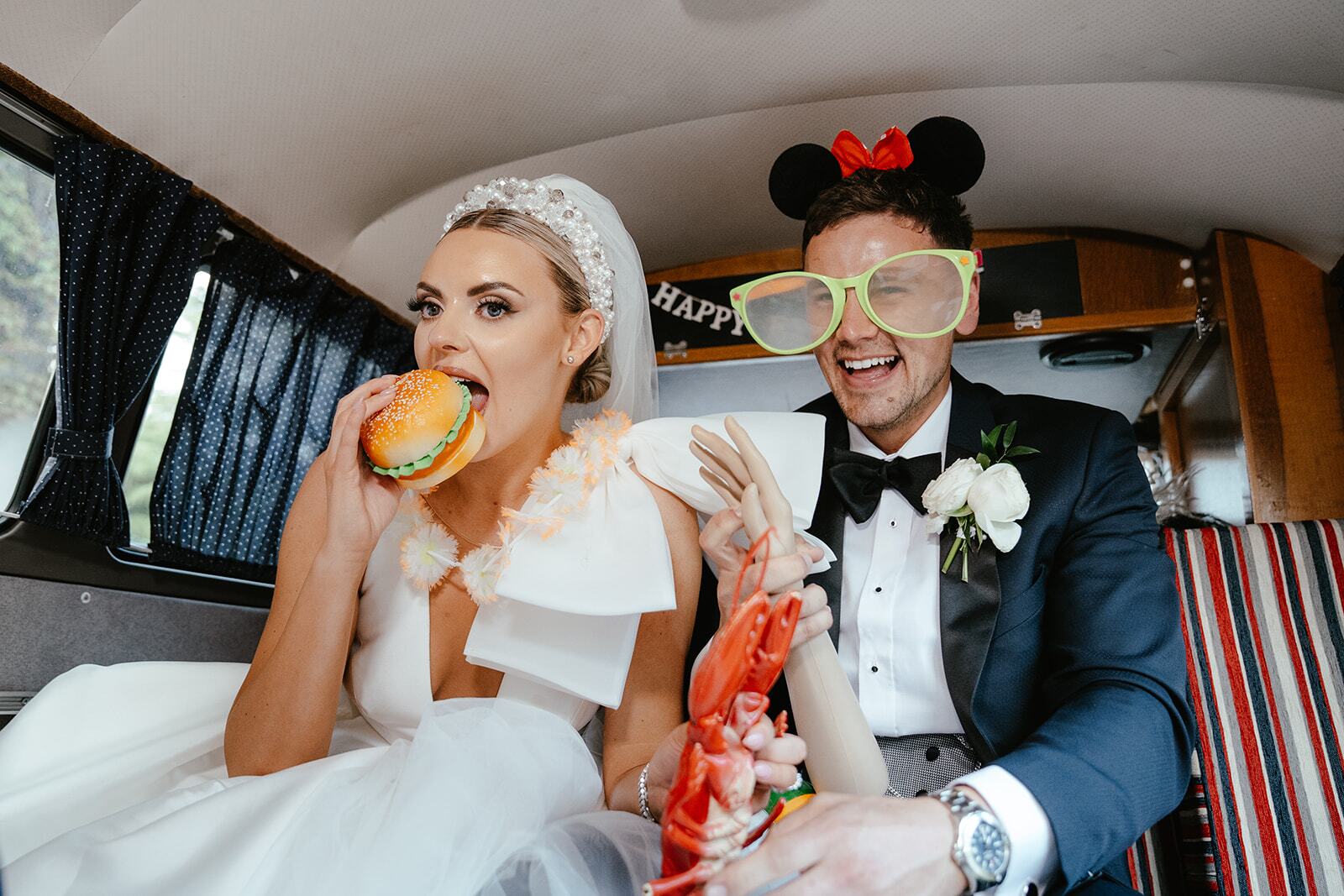 A bride and groom sitting in the back of a van eating a burger.